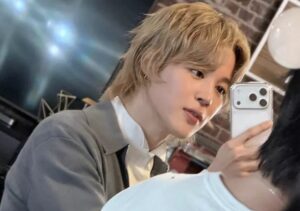 Jimin takes a mirror selfie with a smartphone, dressed in a gray jacket and white shirt in a cozy studio setting.