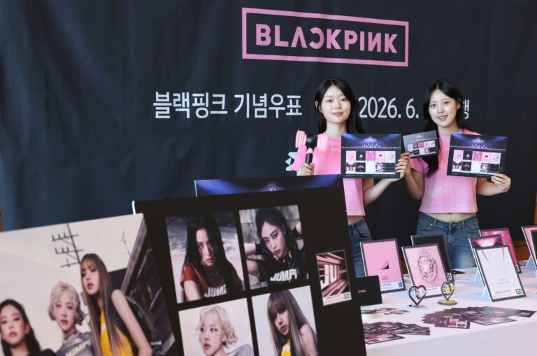 Two models wearing pink shirts display BLACKPINK commemorative stamp sheets at a promotional event. The backdrop features the BLACKPINK logo and Korean text announcing the stamp release date. The table in front of them is filled with BLACKPINK-themed merchandise, including stamp designs, posters, and light sticks.