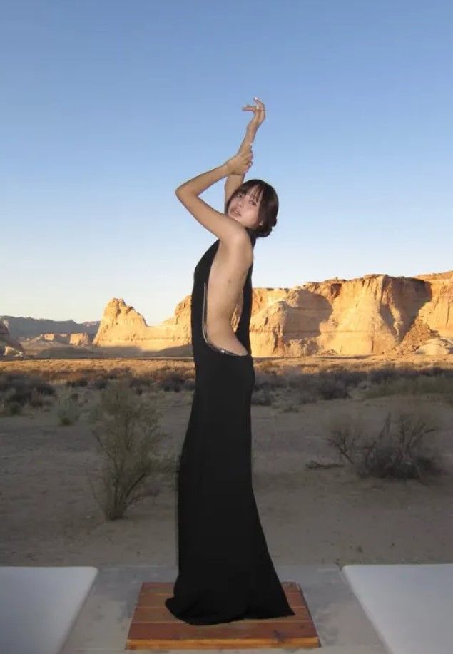 BLACKPINK’s Lisa poses in a sleek, backless black gown with gold detailing, captured in minimalist interiors and a desert landscape at sunset. Her elegant silhouette contrasts against stone walls and glowing cliffs.