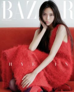 NewJeans’ Hyein in a red dress and shoes sitting on a red couch against a gray background, Harper’s Bazaar Korea September 2023 cover.