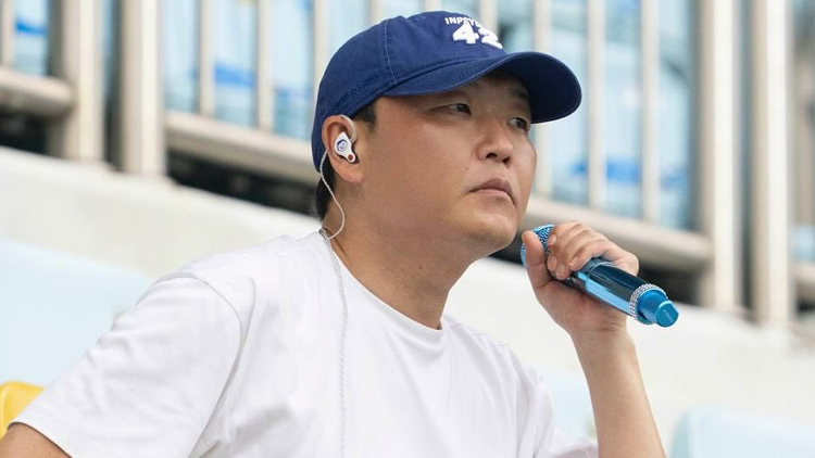 Korean singer PSY wearing a blue baseball cap with the number “42,” a white shirt, and an in-ear monitor, holding a blue microphone while seated in a stadium with light blue seats and railings in the background.