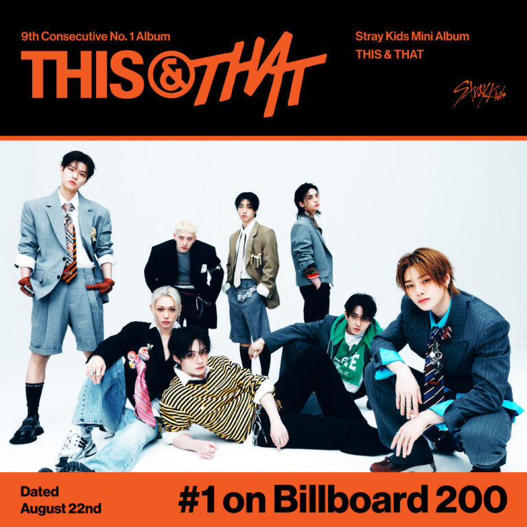 Group photo of Stray Kids in coordinated black suits, standing in front of a turquoise background with bold “THIS & THAT” text, each member raising one hand in greeting.